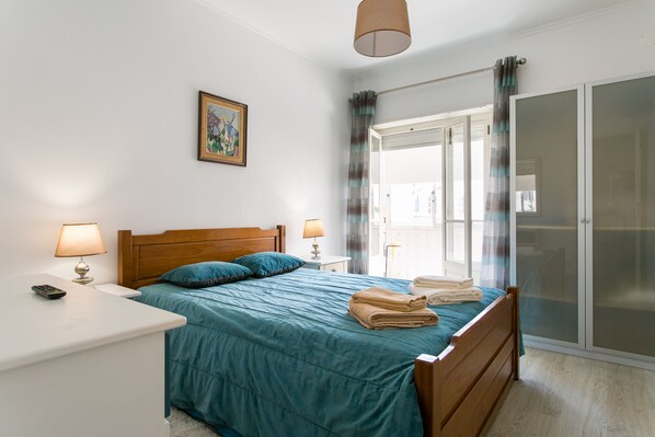 A bedroom with a bed, two bedside tables, a nightstand, a lamp, a painting on the wall, and a sliding glass door with curtains.