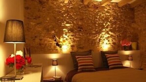 Un dormitorio con pared de piedra, cama con almohadillas a rayas, mesita de noche con lámpara y un jarrón con flores rojas.