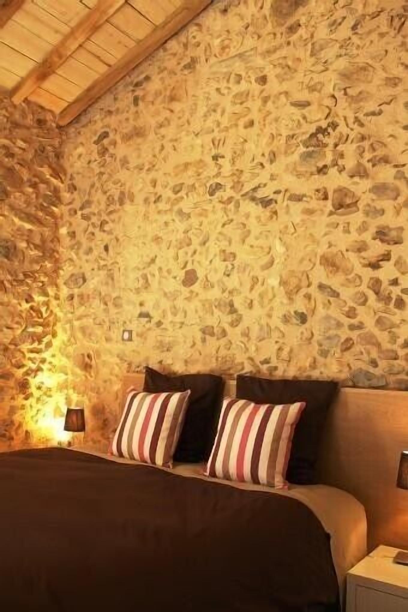 A bedroom with a stone wall, a bed with striped pillows, and a nightstand with a lamp.