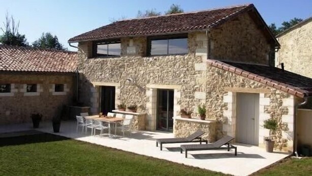 Un edificio de piedra con techo de tejas, un patio con sillas y una mesa, y un pequeño jardín.