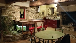 Een rustieke stenen keuken met een ronde tafel en rode stoelen, een stenen open haard en een klein raam met gordijn.