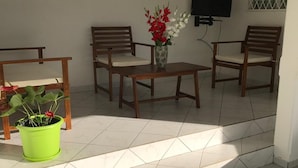 Un patio soleado con suelo de baldosas, dos sillas de madera, una mesita, una planta en maceta y un televisor montado en la pared.