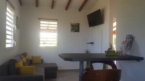 A living room with a sofa, a dining table, a TV mounted on the wall, and a window with blinds. - COZY COTTAGE + NETFLIX + WIFI + JACCUZZI + PARKING (Sainte rose)