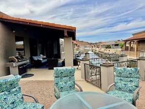 Un espacio al aire libre cubierto con sillas y una mesa, una parrilla, y vista a un canal con casas al fondo.