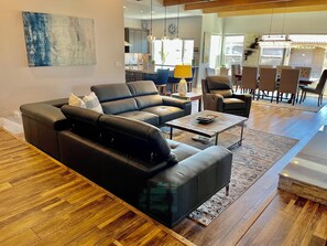 A modern living room with a black leather sofa, a coffee table, and a dining area with chairs and a chandelier.