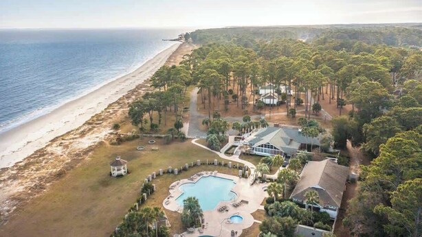An aerial view of a coastal property with a swimming pool, beach access, and a large house surrounded by trees.