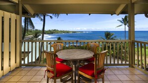 A covered outdoor seating area with a wooden table and chairs, overlooking a beach with palm trees and the ocean.
