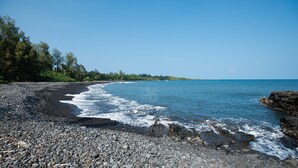 A beach with a pebbly shore, clear blue water, and a line of trees in the background.