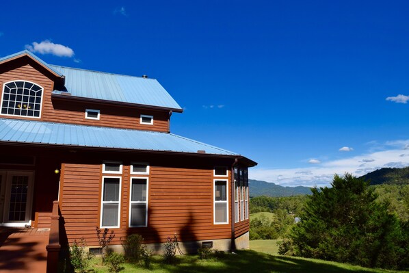 Una casa de dos pisos con fachada marrón y techo azul, rodeada de abundante vegetación y montañas al fondo.