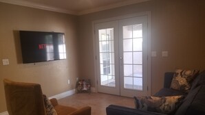 A living room with a TV mounted on the wall, a brown armchair, a blue sofa, and a sliding glass door leading to an outdoor area. - CLOSE TO STRIP Very Private, GATED, Huge Open Floor Plan, WIFI, WASHER & DRYER (Las Vegas)