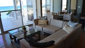 A modern living room with a beige sofa, glass coffee table, and a view of the ocean through large windows. - Early Holiday Booking Discount! (Tulum)