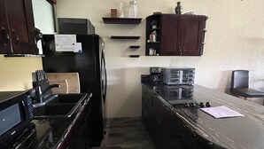 Een kleine keuken met donkere kastjes, een zwart aanrecht, een koelkast, een magnetron, een koffiezetapparaat en een klein plankje met potten.