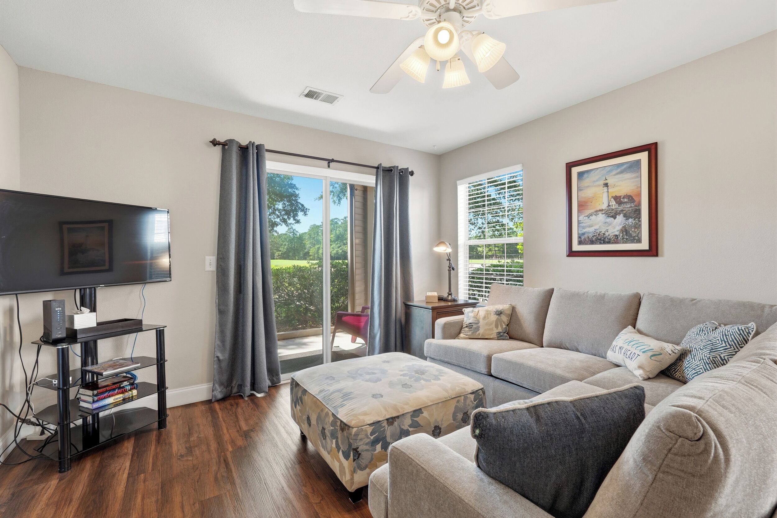 A living room with a flat-screen TV, a sofa, an ottoman, and a ceiling fan.