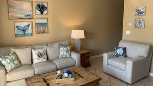 Una sala de estar con un sofá gris, un sillón a juego, una mesa de centro de madera y una lámpara. Hay cuadros colgados en la pared y un enchufe.