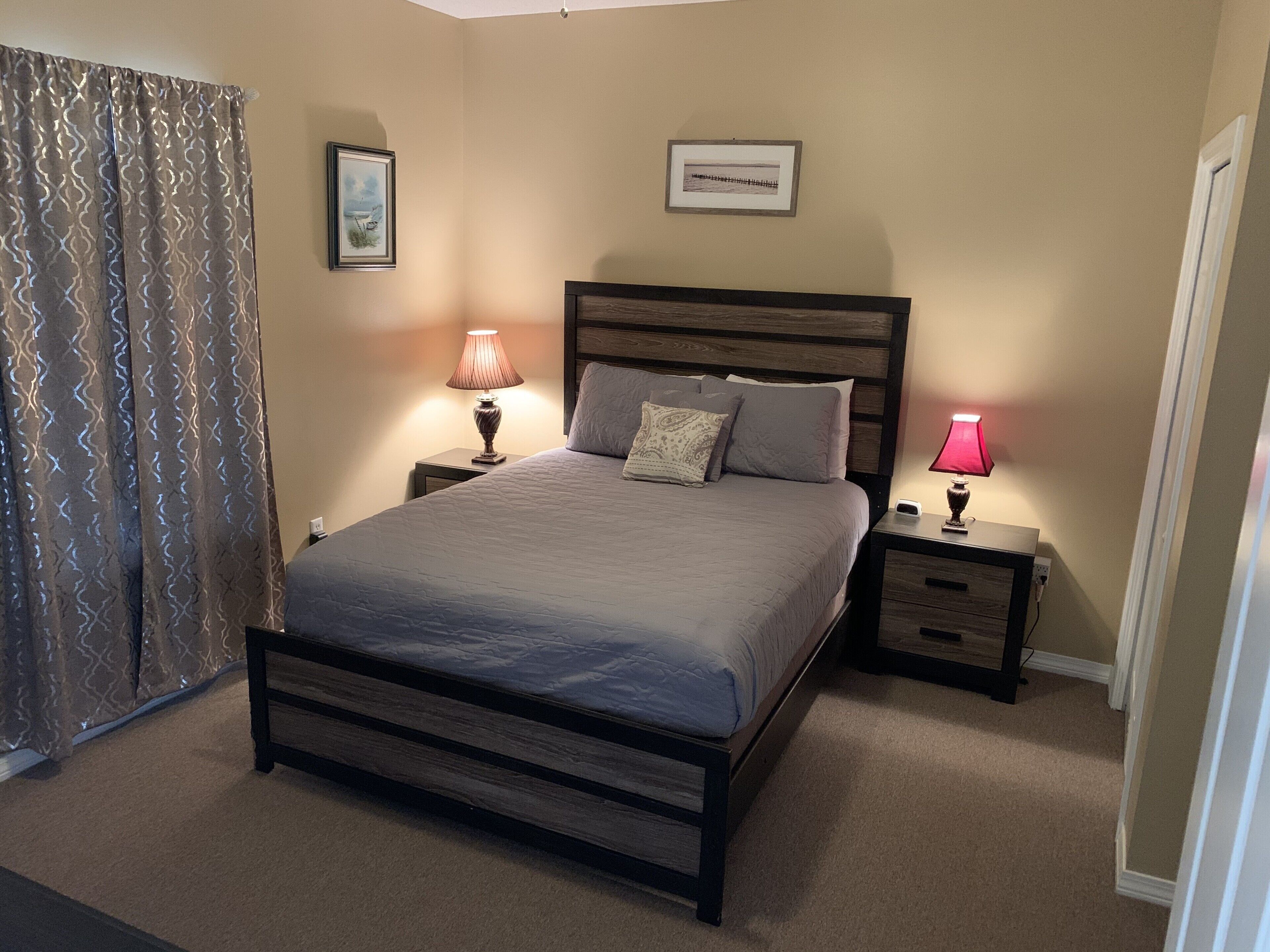 Une chambre à coucher avec un lit, deux tables de chevet, une lampe, un tableau encadré et un rideau.