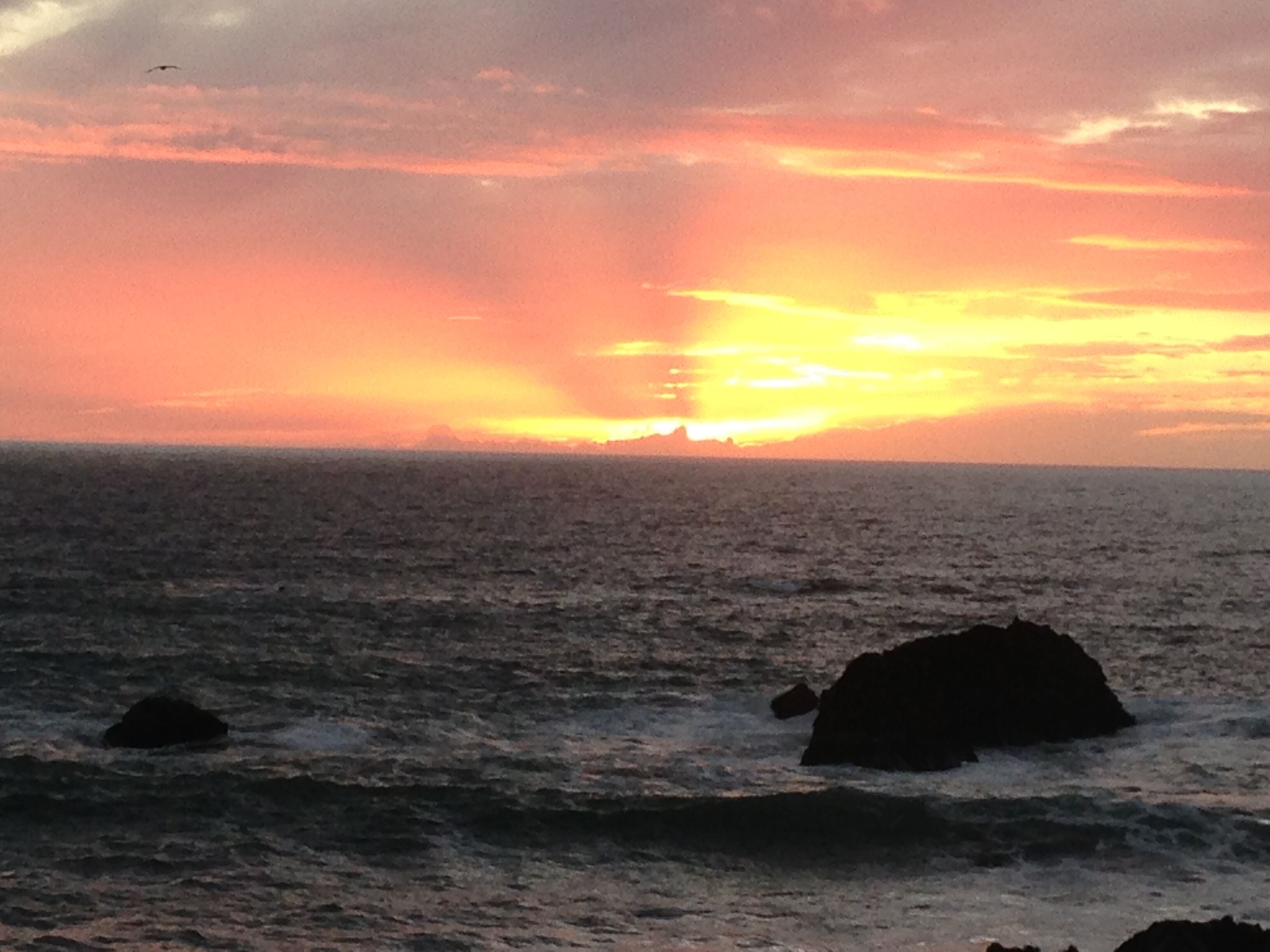 Un coucher de soleil sur l’océan, avec des vagues et des rochers.