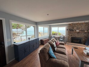 A living room with a view of the sea, a stone fireplace, and a wooden dining table. - Best views on Sonoma Coast!Oceanfront,private hot tub, fire pit, pets considered (Bodega Bay)