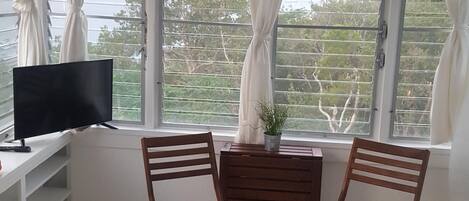 Une pièce avec un téléviseur posé sur une étagère blanche, deux chaises en bois, une petite table en bois et une vue sur le paysage urbain, visible à travers de grandes fenêtres ornées de rideaux blancs.