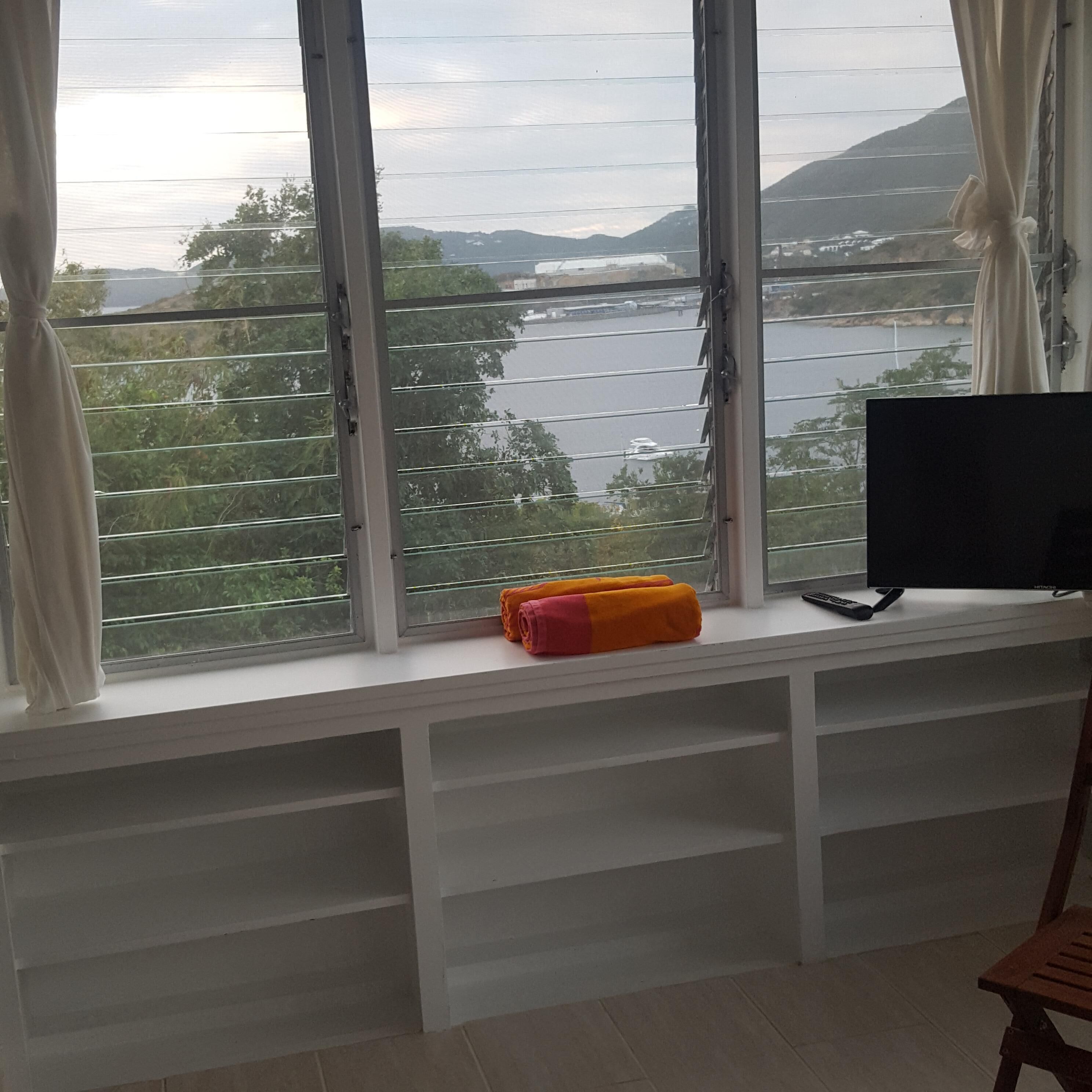 Ein Zimmer mit einem großen Fenster mit Blick auf ein Gewässer und Hügel. Auf der Fensterbank liegt ein zusammenrolltes Handtuch und neben einem Flachbildfernseher liegt eine Fernbedienung.