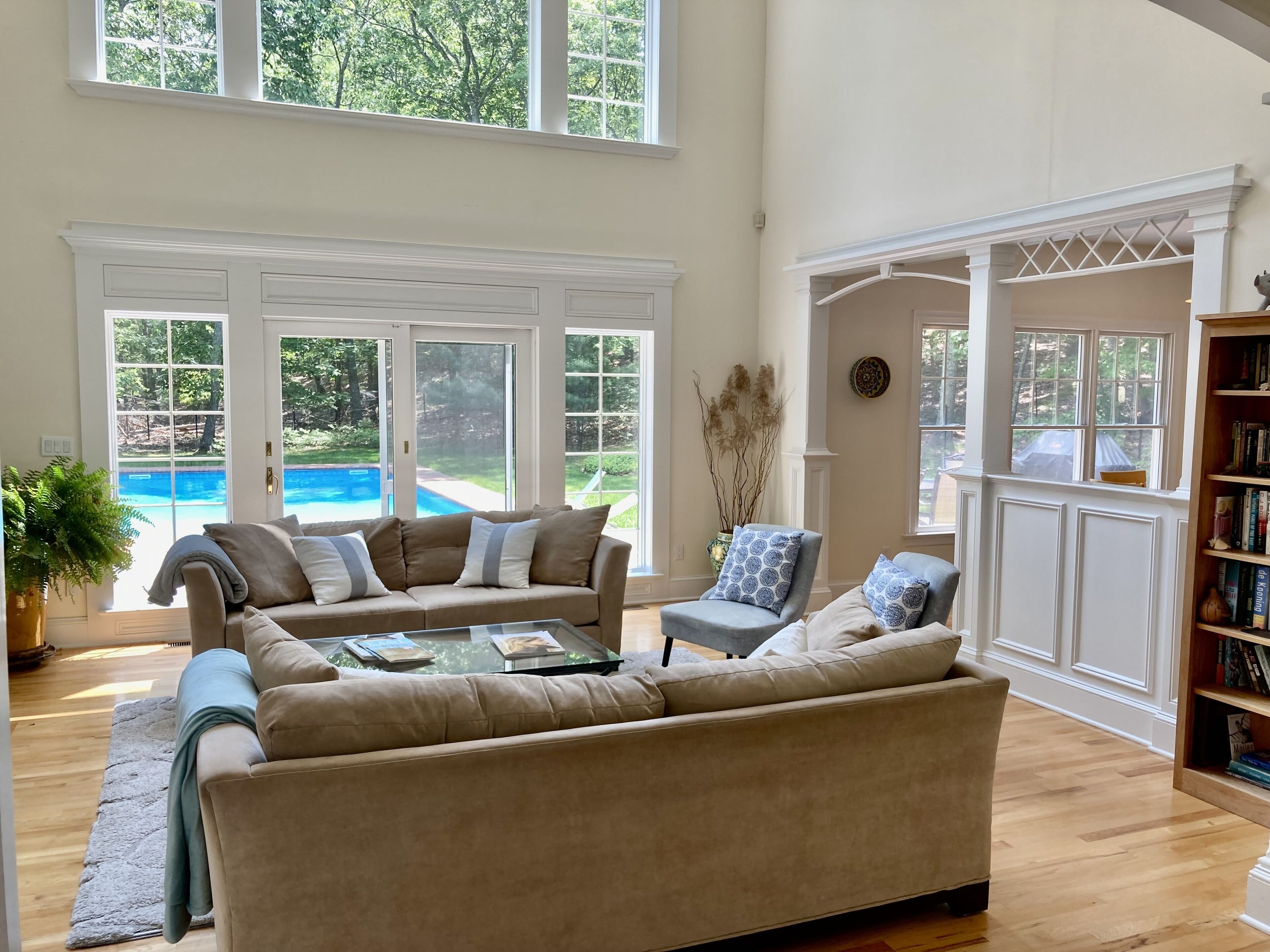 Un salón espacioso con un sofá seccional color beige, una mesa de centro de cristal y una estantería. Amplias ventanas ofrecen vistas a un jardín trasero con piscina.