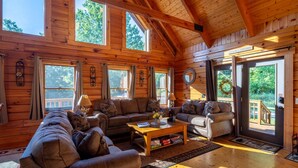 Un intérieur spacieux de chalet en bois, avec plusieurs coins salon, de grandes fenêtres et une vue sur l’extérieur.