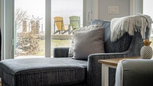 A modern living room with a grey sofa, a small wooden side table, and a view of outdoor chairs through a large window. - Another Sunset by the Bay: Waterfront - 2-Bedroom (Leamington)