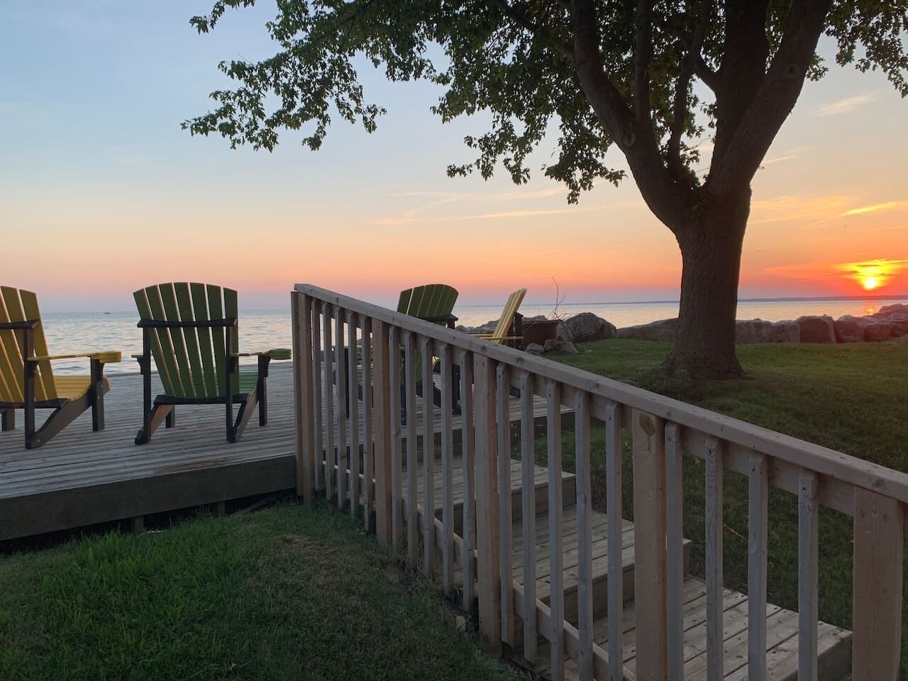 Une terrasse en bois avec des fauteuils Adirondack, offrant une vue sur le coucher de soleil sur l’eau.
