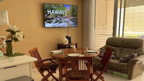 A living room with a dining area, a television mounted on the wall, a ceiling fan, and a window with blinds.