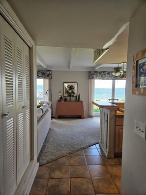 A room with a sofa, a small table with decorative items, a window with a view of the sea, and a kitchen area with cabinets.