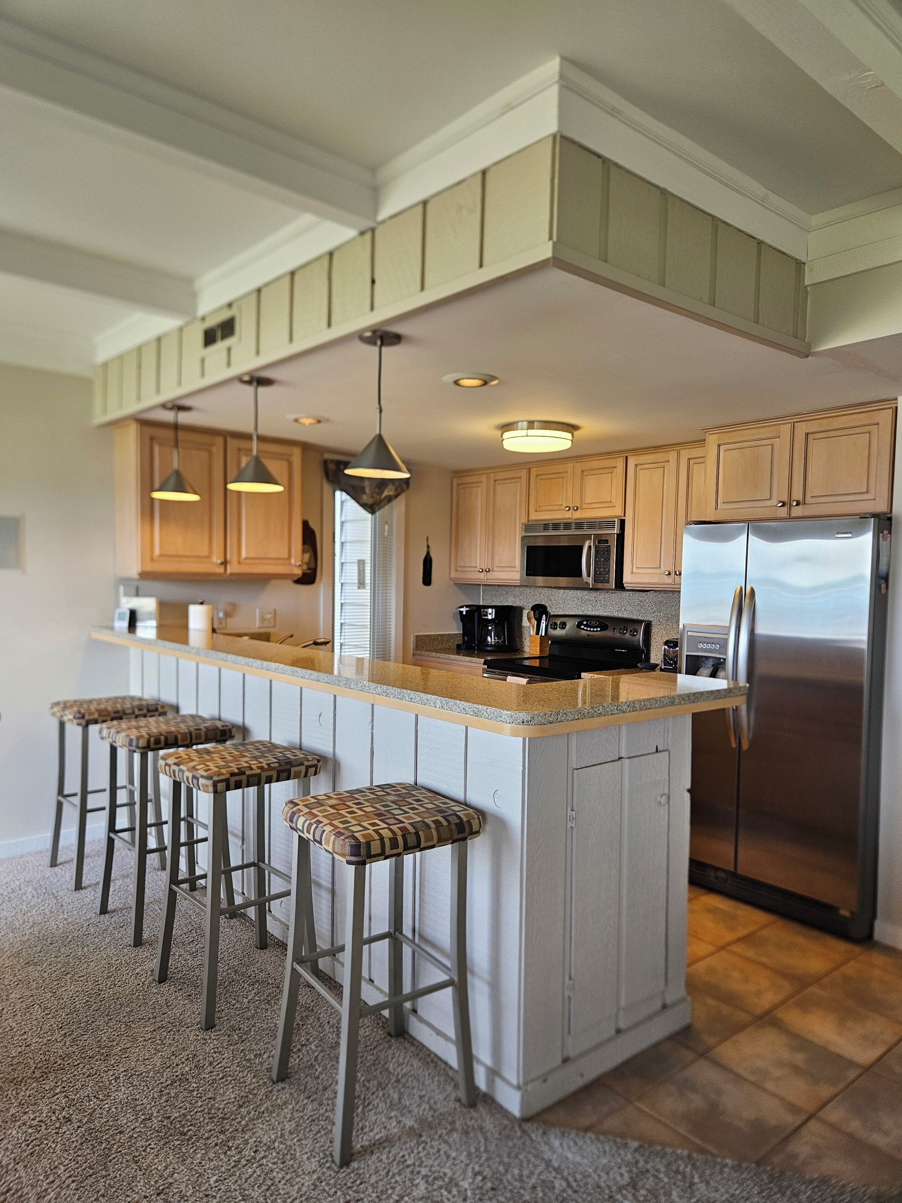 A kitchen with a central island, bar stools, wooden cabinets, stainless steel appliances, pendant lighting, and a tiled floor.