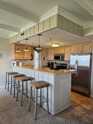 A kitchen with a central island, bar stools, wooden cabinets, stainless steel appliances, pendant lighting, and a tiled floor.