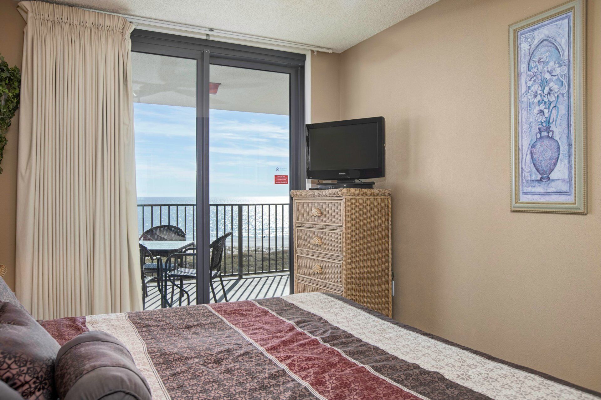 Une chambre d’hôtel avec un lit, un téléviseur posé sur un meuble, une table d’appoint en osier et un balcon équipé d’une table et de chaises.