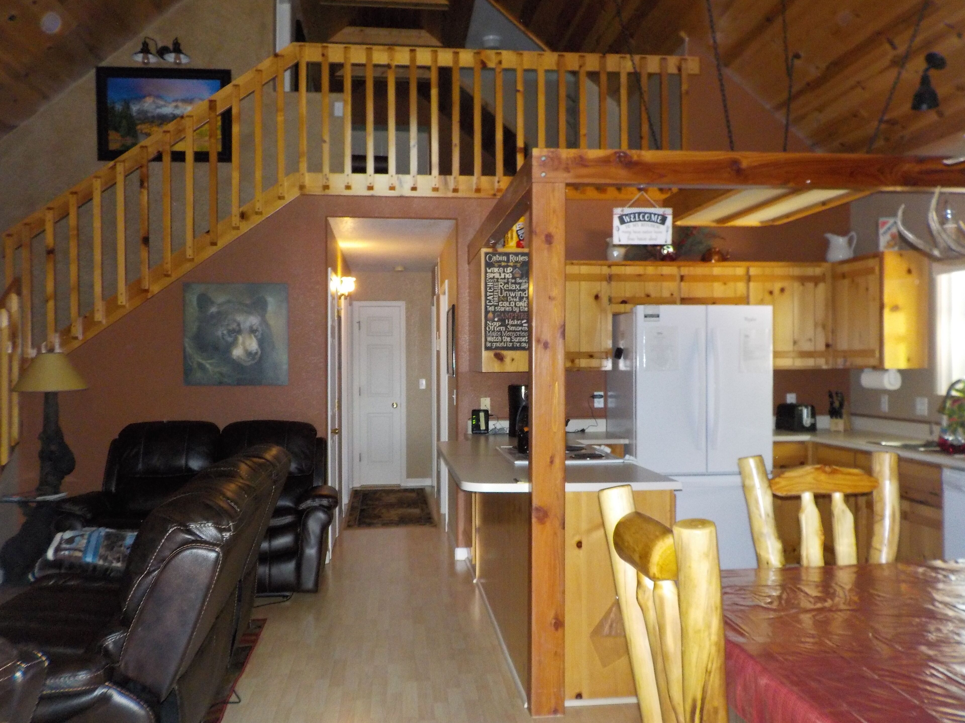 A cabin interior with a wooden staircase, a living area with leather furniture, a kitchen with wooden cabinets, and a dining area with a table and chairs.