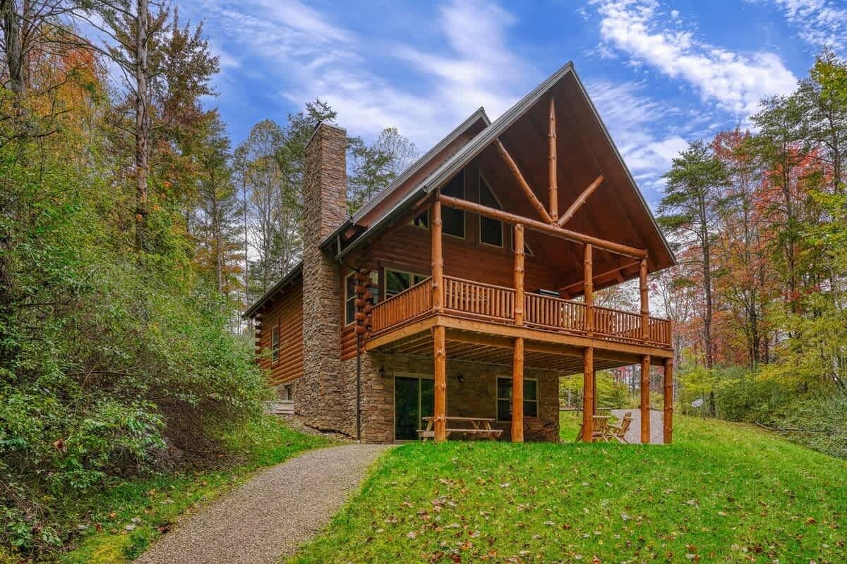 A log cabin with a balcony, surrounded by trees and a gravel path.