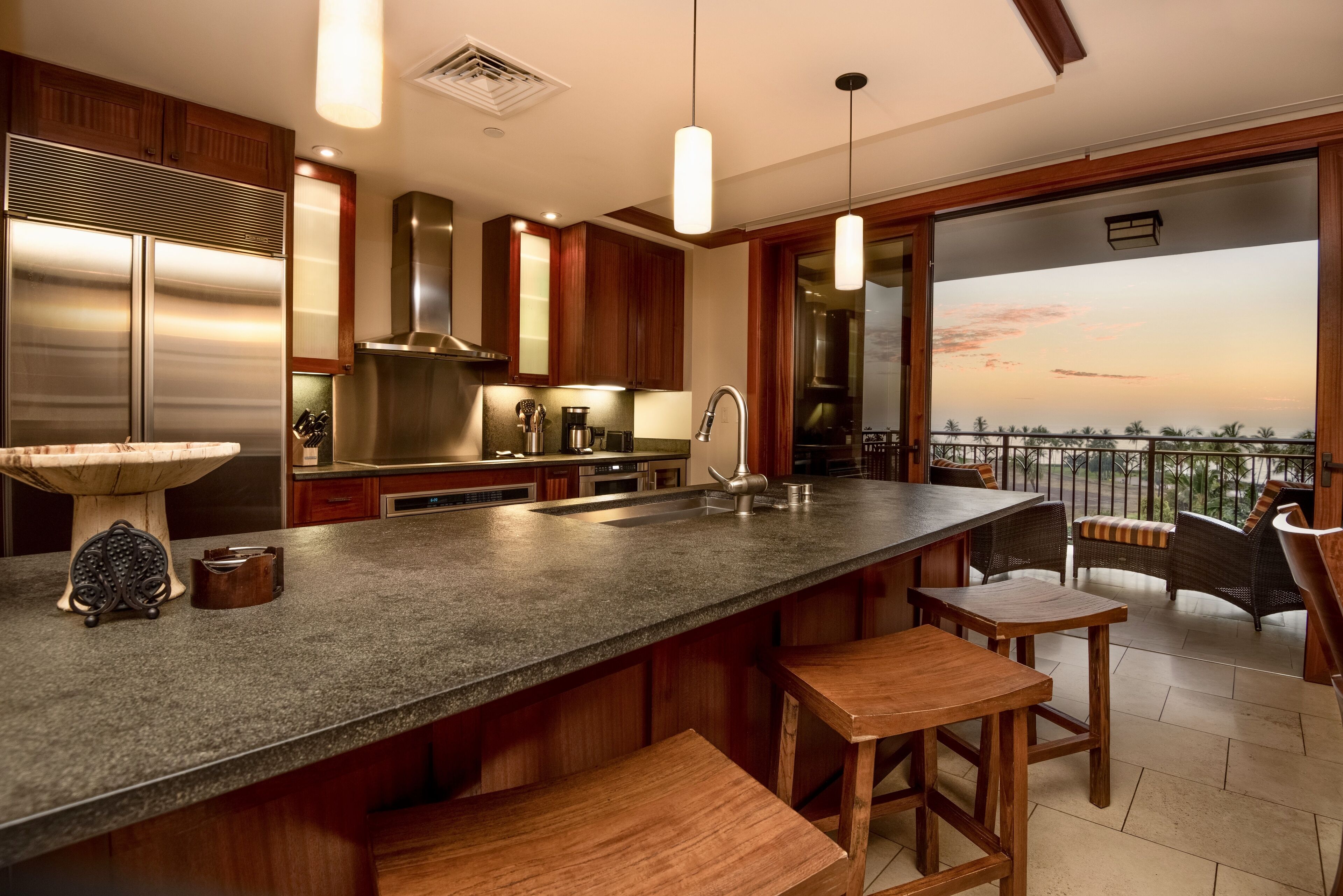 A modern kitchen with a large island, stainless steel appliances, and a view of the sunset from the balcony.