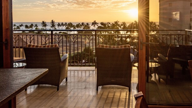 Uma varanda com móveis de vime, vista para o oceano e um pôr do sol.