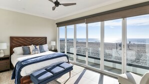 Ein Schlafzimmer mit einem großen Bett, einer Bank, einem Deckenventilator und Blick auf den Strand durch große Fenster.