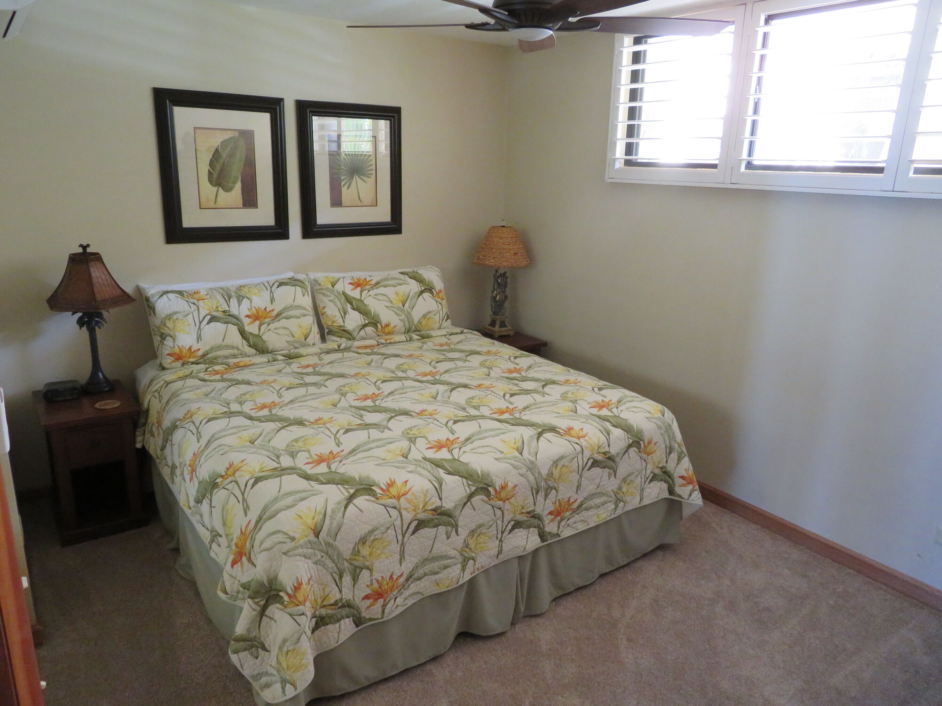 A bedroom with a bed, two framed botanical prints on the wall, a ceiling fan, a window with blinds, and a nightstand with a lamp.