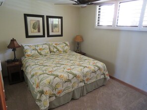 A bedroom with a bed, two framed botanical prints on the wall, a ceiling fan, a window with blinds, and a nightstand with a lamp.