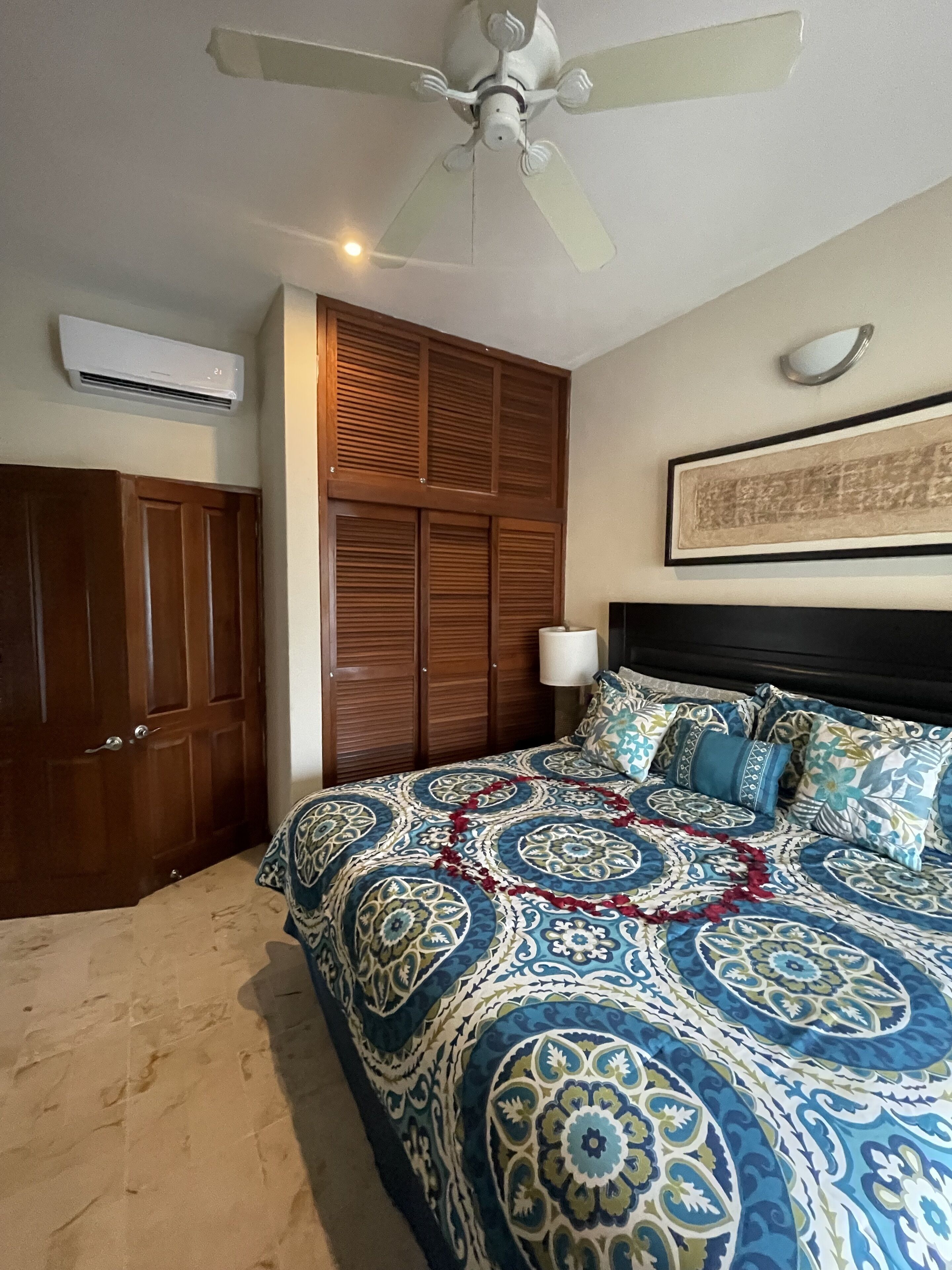 A bedroom with a bed, a ceiling fan, a wooden wardrobe, and an air conditioning unit.