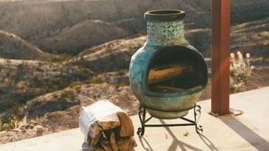 Un entorno rústico al aire libre con un soporte de metal que sostiene una maceta de cerámica, una pila de leña y una estructura de pérgola de metal.