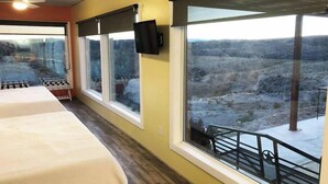 Habitación de hotel con cama, ventilador de techo, televisor montado en la pared y ventanas amplias que ofrecen vistas de un paisaje rocoso.
