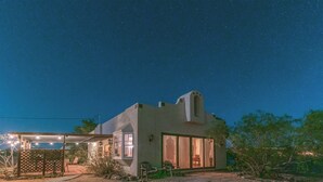 A modern house with a large glass door, a covered patio with chairs, and a starry night sky.