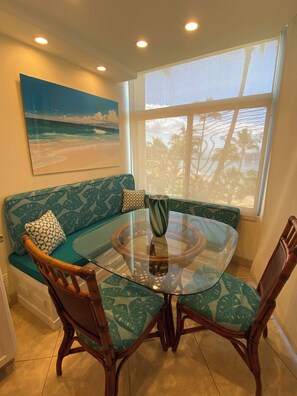 Un comedor con una mesa de vidrio rodeada de sillas de madera con cojines estampados de hojas tropicales, un sofá azul aguamarina con cojines decorativos y un cuadro de una escena de playa en la pared.