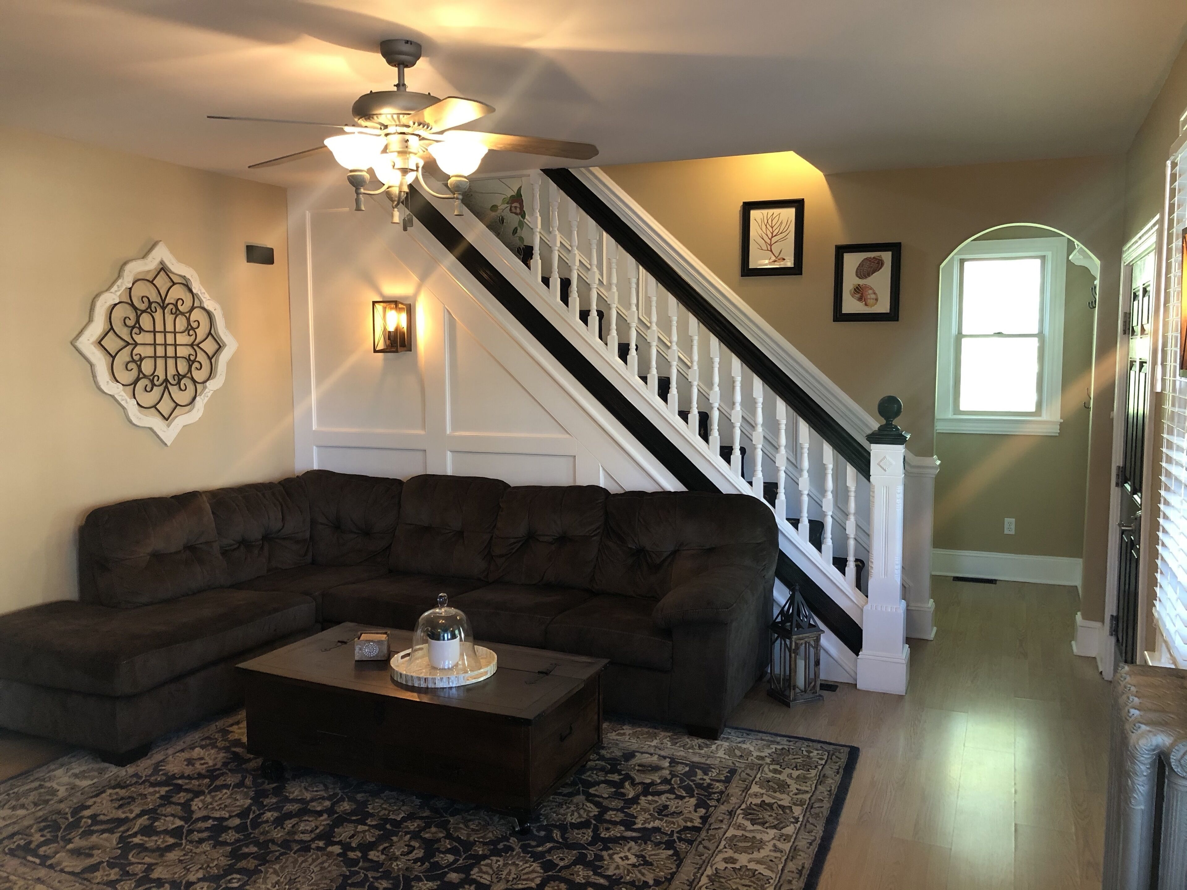 A living room with a dark brown sectional sofa, a wooden coffee table, a patterned rug, a ceiling fan, and a staircase with white railings.