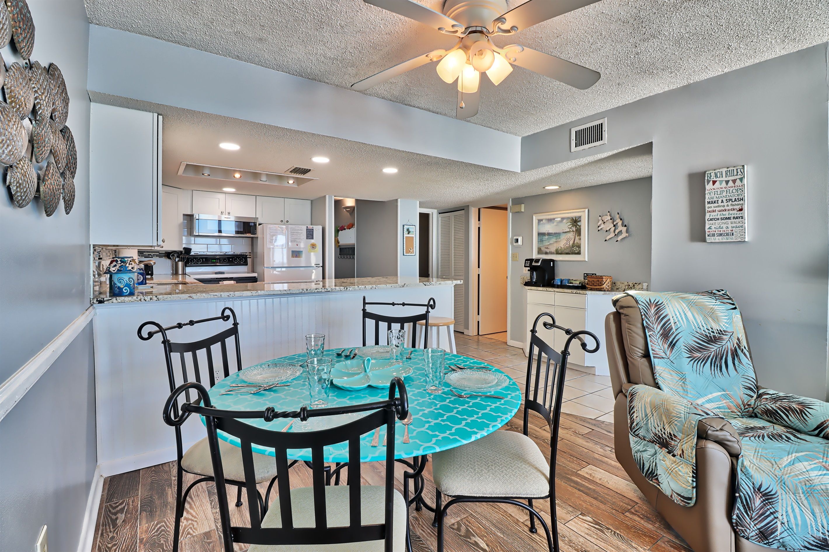 A modern kitchen with white cabinets, a dining area with a turquoise table, and a ceiling fan.