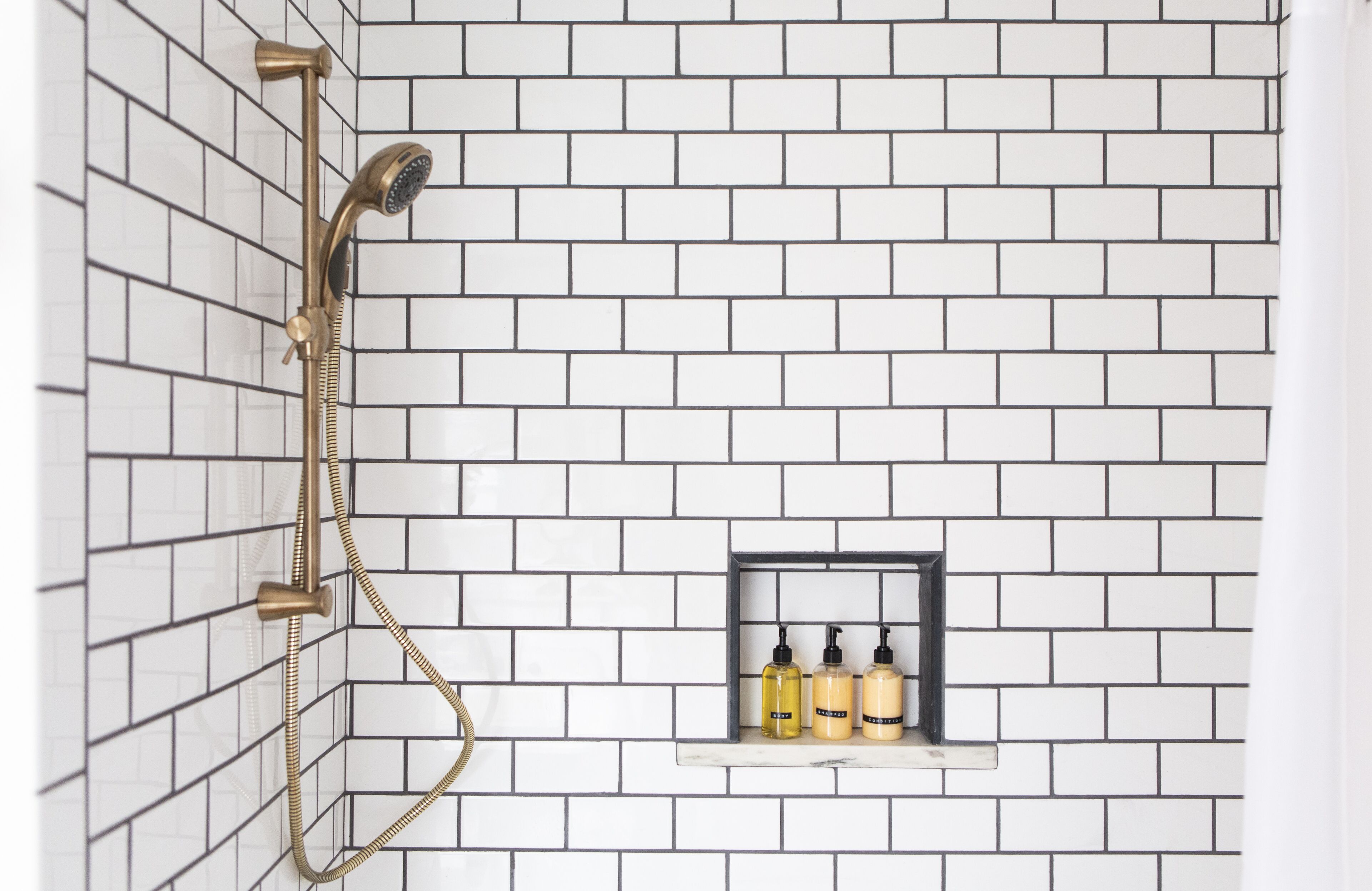A shower with a gold showerhead and hose, white tiled walls, and a shelf with toiletries.