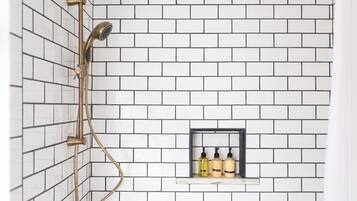 A shower with a gold showerhead and hose, white tiled walls, and a shelf with toiletries.