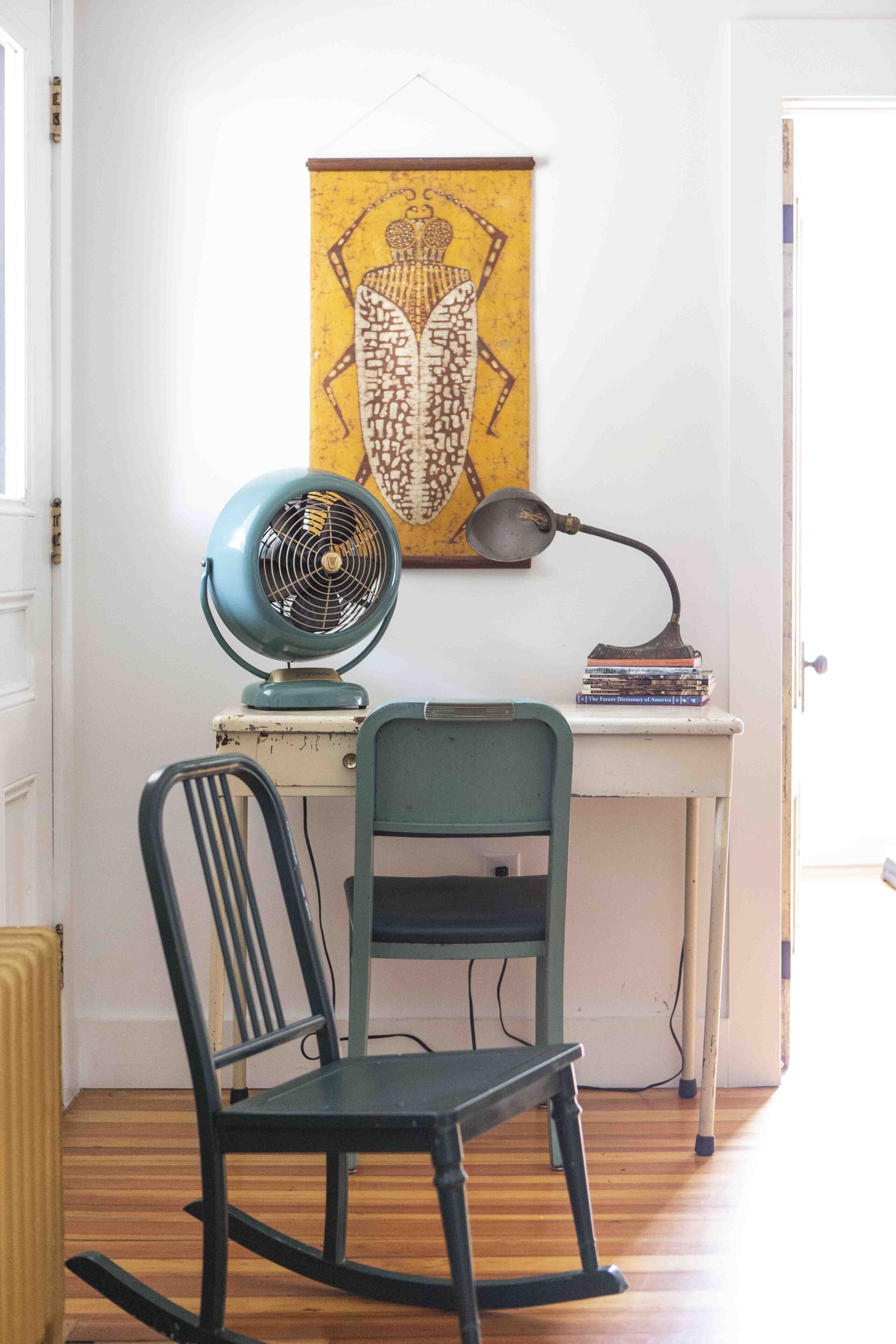 Une pièce avec un plancher en bois, un fauteuil à bascule, un bureau avec une lampe, un ventilateur et une œuvre d’art encadrée accrochée au mur.