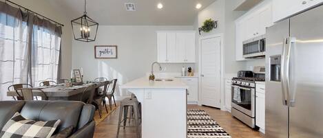 A modern kitchen with white cabinets, a stainless steel refrigerator, and a dining area with a black leather couch and a dining table.